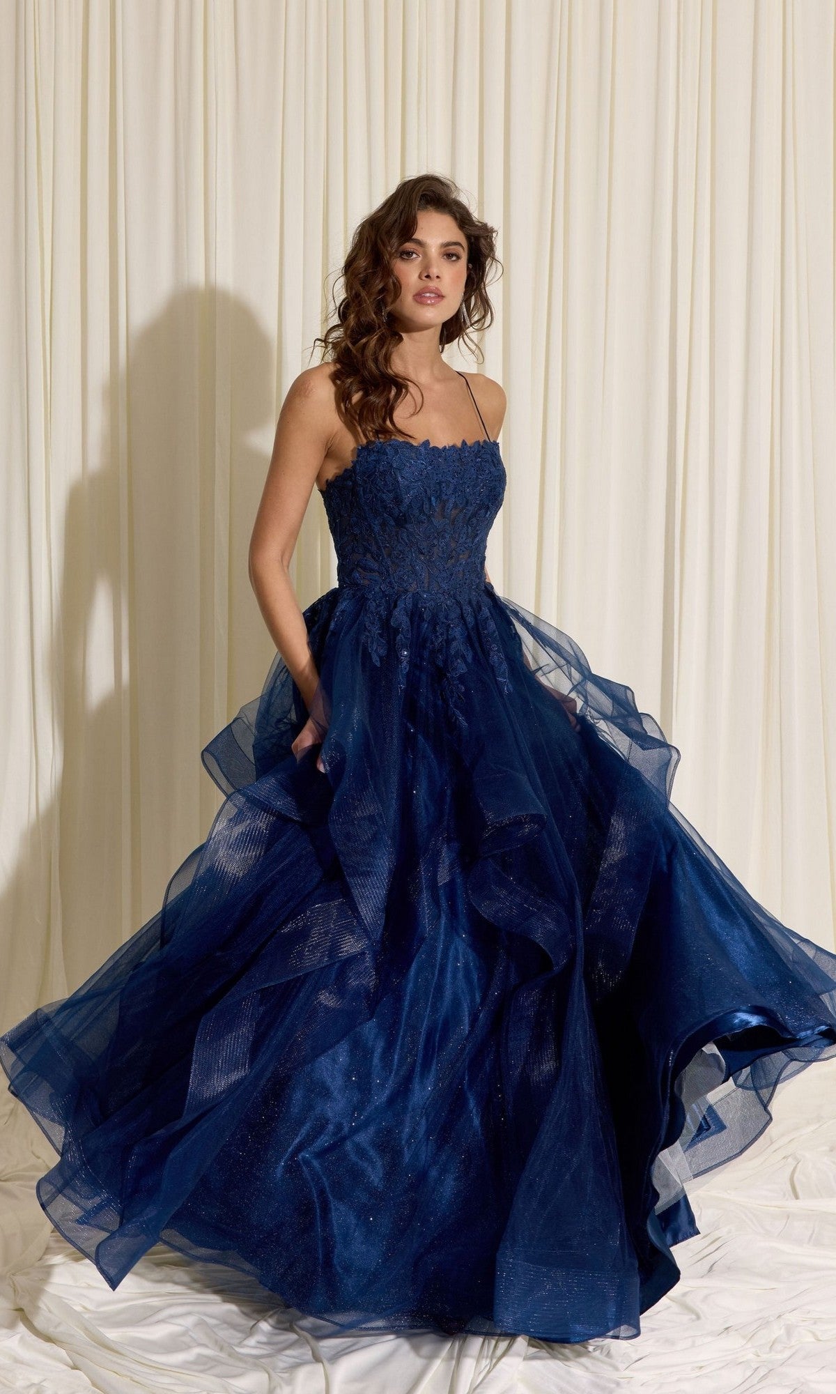 A woman in the Eureka EK167 Long Prom Dress in navy, featuring a strapless floral lace corset and layered tulle A-line skirt, stands before cream draped fabric.
